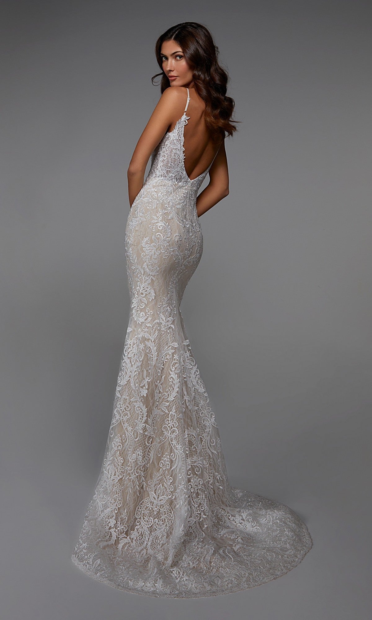 A woman poses in the Alyce 7028 long white dress, a fitted, floor-length evening gown with lace embroidery, a train, and backless design, shown against a plain gray background.
